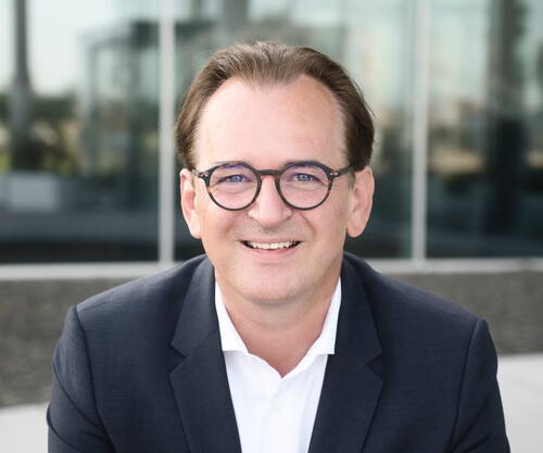 Lächelnder Mann mit Brille im dunklen Anzug und weißem Hemd vor moderner Glasfassade