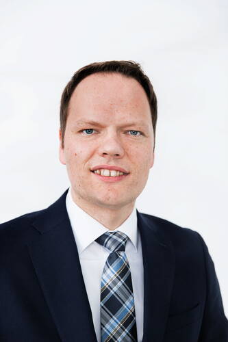 Mann im dunklen Anzug mit kariierter Krawatte lächelt frontal in die Kamera vor hellem Hintergrund