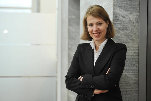 Businessfrau im schwarzen Blazer steht mit verschränkten Armen lächelnd vor einer modernen Bürowand aus Glas und Stein.