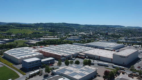 Großes Industrie- und Logistikgelände mit Hallen und Solaranlagen auf den Dächern am Stadtrand, umgeben von grünen Hügeln und Wohnbebauung im Hintergrund