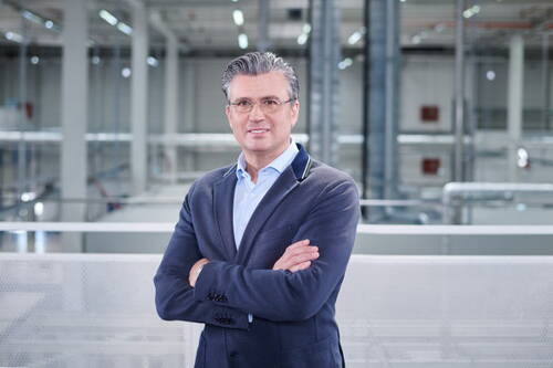 Geschäftsmann mittleren Alters mit Brille und verschränkten Armen steht selbstbewusst in einer modernen Industriehalle mit heller, technischer Umgebung