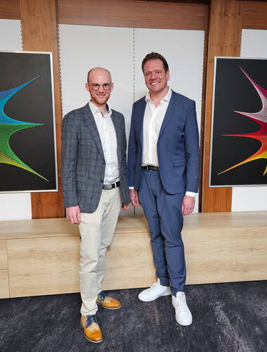 Zwei lächelnde Männer in Businesskleidung stehen nebeneinander in einem modernen Konferenzraum vor einer Holzverkleidung, flankiert von zwei farbenfrohen, abstrakten Wandbildern.