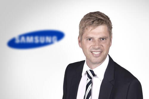 Geschäftsmann im Anzug lächelt in die Kamera, im Hintergrund unscharfes Samsung‑Logo auf hellem Hintergrund