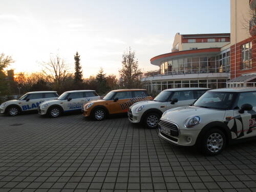 Mehrere gebrandete Mini-Cooper-Firmenwagen stehen in einer Reihe vor einem modernen, verglasten Gebäude bei untergehender Sonne.