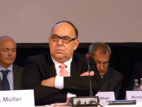 Älterer Mann im Anzug mit verschränkten Armen sitzt ernst an einem Konferenztisch, flankiert von zwei weiteren Männern, die in Unterlagen blicken.