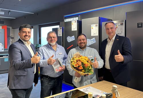 Vier Männer in einem Elektrofachgeschäft lächeln in die Kamera, zeigen den Daumen nach oben und halten als Anerkennung einen Blumenstrauß und ein Dankesgeschenk in der Hand.