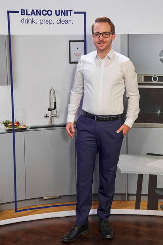 Mann in weißem Hemd und blauer Hose steht lächelnd in einer modernen Küche vor einem BLANCO UNIT Spülbereich mit Hocharmatur und Obstschale auf der Arbeitsplatte