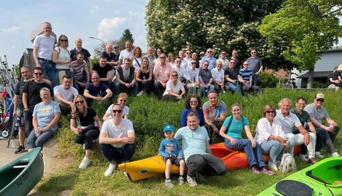 Große Gruppe fröhlicher Menschen sitzt an einem sonnigen Frühlingstag auf einer Wiese vor blühenden Bäumen und bunten Kajaks und posiert gemeinsam in die Kamera.
