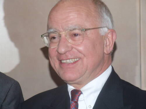 Älterer, lächelnder Mann mit Brille und Glatze in Anzug und roter Krawatte in einem Innenraum.