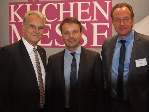 Drei Geschäftsleute in Anzügen posieren lächelnd nebeneinander vor einem pinken Messeschild mit der Aufschrift „Küchenmesse“.