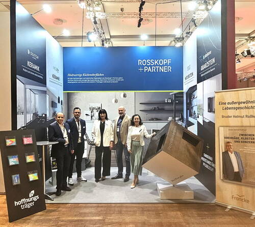 Fünf Geschäftsleute posieren auf einer Messe vor dem Rosskopf+Partner‑Messestand mit modernen Küchenoberflächen, Infotafeln und Werbedisplays.