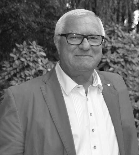 Älterer Mann mit weißem Haar und Brille lächelt in einem Anzugjackett und weißem Hemd vor unscharfem, bewaldetem Hintergrund