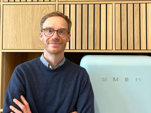 Mann mit Brille und Bart lächelt mit verschränkten Armen vor einer hellblauen SMEG-Kühltruhe und einer Holzschrankwand