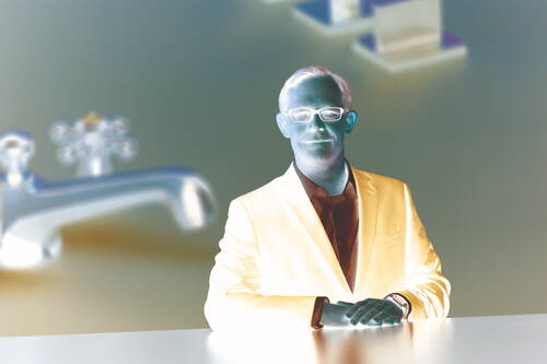 Mittelalter Mann im blauen Anzug lehnt mit gefalteten Händen an einem Tisch vor unscharfem Hintergrund mit vergrößerter goldener Wasserhahn-Installation