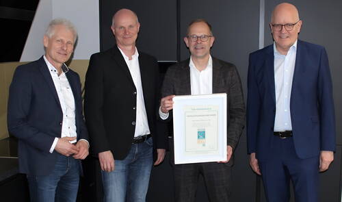 Vier Männer in Businesskleidung stehen nebeneinander in einem Büro, während der zweite von rechts eine gerahmte Verleihungsurkunde in die Kamera hält.