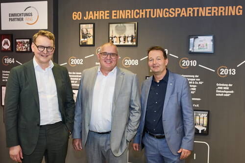 Drei lächelnde Männer im Anzug stehen vor einer bebilderten Zeitleiste zum 60-jährigen Jubiläum des Einrichtungspartnerrings.