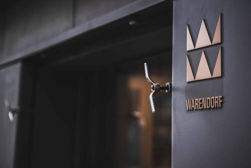 Eingangstür mit Metallgriff und modernem WARENDORF-Logo aus goldfarbenen Dreiecken auf dunkler Fassade