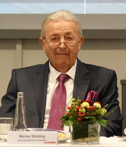 Älterer Herr im dunklen Anzug mit pinker Krawatte sitzt an einem Konferenztisch mit Namensschild, Wasserglas und kleinem Blumenarrangement vor sich.