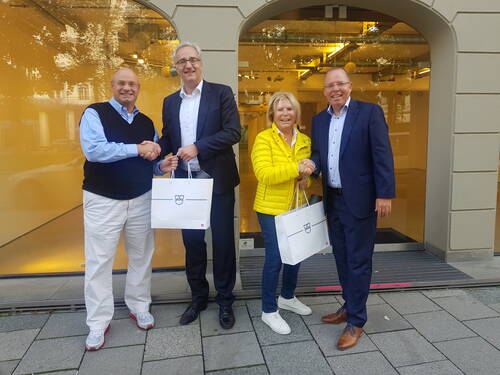 Vier lächelnde Personen stehen vor einem Schaufenster, schütteln sich paarweise die Hände und halten weiße Geschenktaschen mit demselben Logo.