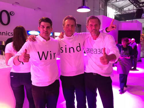 Drei Männer in weißen T-Shirts mit dem Aufdruck „Wir sind area30“ stehen arm in arm auf einer beleuchteten Messeveranstaltung und zeigen begeistert den Daumen hoch.