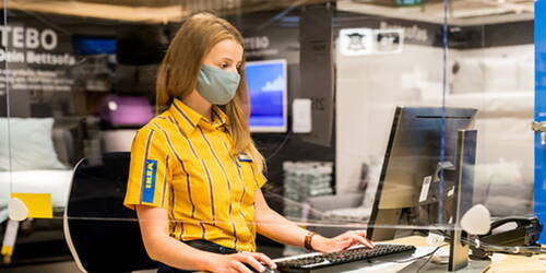 Mitarbeiterin im gelb-blau gestreiften IKEA-Hemd arbeitet mit Mund-Nasen-Schutz an einem Computer hinter einer Plexiglasscheibe im Einrichtungshaus