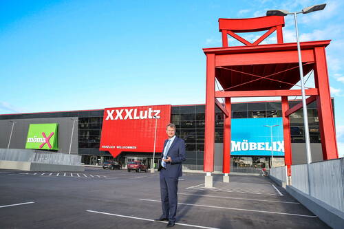 Geschäftsmann im Anzug steht auf einem leeren Parkplatz vor einem modernen Möbelhauskomplex mit riesigem roten Stuhl und den bunten Fassaden von XXXLutz, mömax und Möbelix