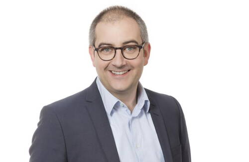 Lächelnder Mann mit Brille in Hemd und dunklem Anzug vor weißem Hintergrund