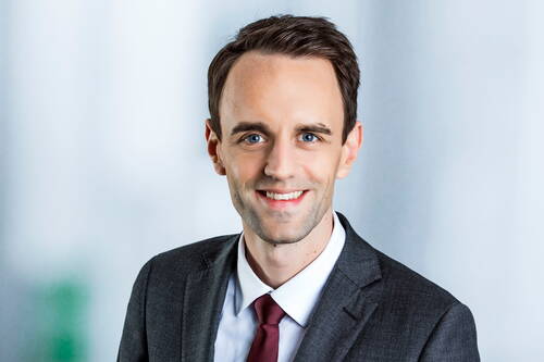 Junger Geschäftsmann im grauen Anzug mit dunkelroter Krawatte lächelt vor neutralem, hellgrauem Hintergrund in die Kamera.