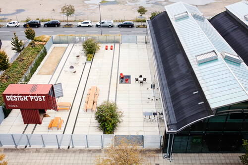 Modern gestalteter Innenhof mit Container „Design Post“, langen Holzbänken, vereinzelten Sitzgruppen und angrenzender Halle mit gewölbtem Glasdach in urbaner Umgebung