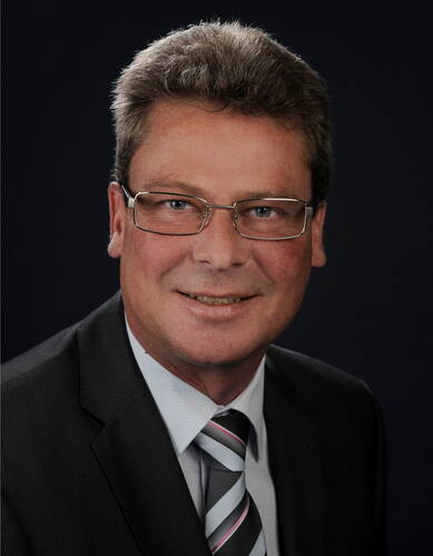 Mittelälterer Mann mit Brille im dunklen Anzug und gestreifter Krawatte lächelt vor dunklem Hintergrund in die Kamera