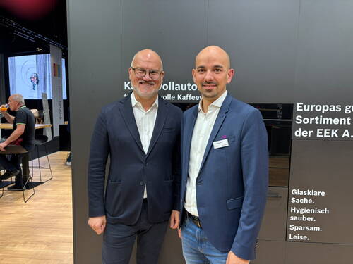 Zwei lächelnde Männer in Anzügen stehen nebeneinander vor einem Messestand mit dunkler Wand und weißer Beschriftung, während im Hintergrund Besucher an Stehtischen sitzen.