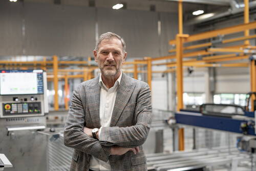 Geschäftsmann im karierten Sakko steht mit verschränkten Armen in moderner Industriehalle vor einer CNC-Maschine