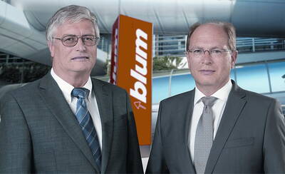 Zwei Geschäftsführer in Anzügen posieren vor einem modernen Firmengebäude mit orangefarbenem Blum-Logo im Hintergrund