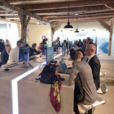 Teilnehmer eines Workshops arbeiten in einem modernen, offen gestalteten Büro gemeinsam an Computern entlang einer langen Tischreihe.
