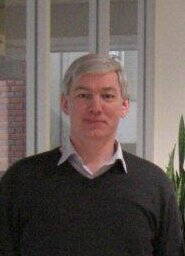 Mittelalterlicher Mann mit grauen Haaren und Hemd unter dunklem Pullover steht in einem hellen Innenraum vor Glaswänden und einer Zimmerpflanze.