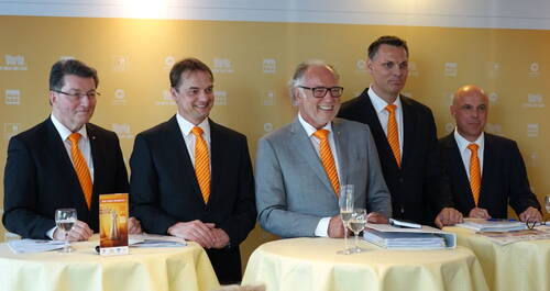 Fünf geschäftlich gekleidete Männer mit orangefarbenen Krawatten stehen lächelnd an Stehtischen mit Unterlagen und Gläsern vor einer gelb gebrandeten Pressewand.