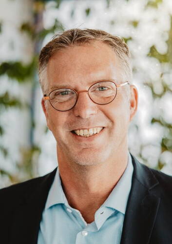 Lächelnder Mann mittleren Alters mit Brille und grauem Haar in Businesskleidung vor unscharfem, grün bewachsenem Hintergrund