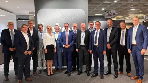 Gruppe von Geschäftsleuten in Anzügen und Businesskleidung posiert lächelnd in einer modernen Messe- oder Ausstellungsumgebung vor einer großen, grauen Wand mit Firmenlogo.