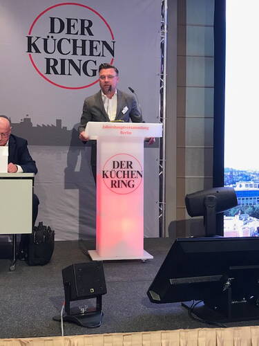 Redner im grauen Anzug spricht am beleuchteten Rednerpult der Jahreshauptversammlung von „Der Küchenring“ in Berlin vor großer Leinwand und Publikum