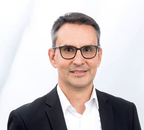 Mittelalterlicher Mann mit Brille im dunklen Anzug und weißem Hemd lächelt dezent vor hellem Hintergrund