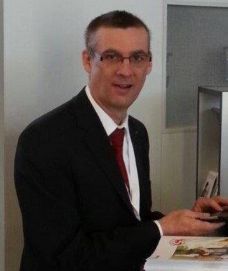 Mann im Anzug mit roter Krawatte lächelt in die Kamera, während er an einem Schreibtisch auf ein Smartphone schaut