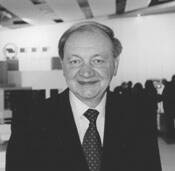 Älterer Mann im Anzug lächelt in einer modernen, schwarz-weiß aufgenommenen Ausstellungs- oder Messehalle in die Kamera