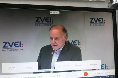 Mann im Anzug spricht mit eingeschaltetem Mikrofon während einer Online-Konferenz vor einer ZVEI-Wand mit eingeblendeten Teilnehmernamen auf dem Bildschirm