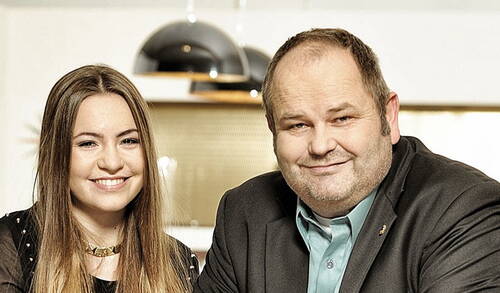 Junge Frau und älterer Mann sitzen lächelnd nebeneinander in einem modern eingerichteten, warm beleuchteten Raum