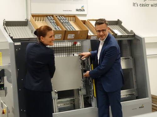 Zwei Geschäftsleute in Anzugkleidung demonstrieren gemeinsam eine industrielle Prüfmaschine für Möbelauszüge in einem Ausstellungsraum von Hettich.
