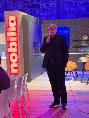 Mann im schwarzen Anzug spricht mit Mikrofon in modern beleuchtetem Showroom mit Nobilia-Leuchtschild und eingerichteter Küchenausstellung im Hintergrund.