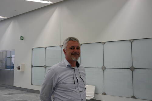 Mittelalterlicher Mann mit grauen Haaren und Bart lächelt in einem modernen, hellen Büroflur mit großen Milchglaswänden im Hintergrund