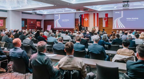 Großer Konferenzsaal mit dicht besetztem Publikum, das einem Vortragenden auf der Bühne vor zwei Leinwänden und mehreren Landesflaggen aufmerksam zuhört