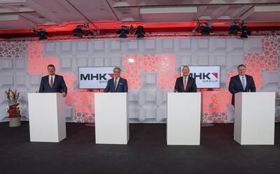 Vier Manager im Anzug stehen hinter Rednerpulten vor einer beleuchteten Bühnenwand mit MHK-Group-Logos während einer Unternehmensveranstaltung