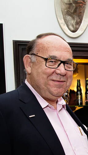 Älterer Mann mit Brille und Anzugjacke lächelt in einer Bar, im Hintergrund stehen mehrere Flaschen auf einem Regal.